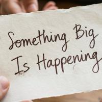 Hands holding a handwritten note that says "Something Big Is Happening"