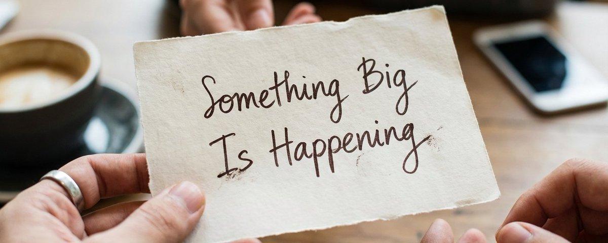 Hands holding a handwritten note that says "Something Big Is Happening"